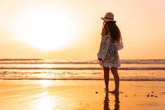 Uma mulher ao pôr do sol em um vestido branco com um chapéu andando à beira-mar na maré baixa