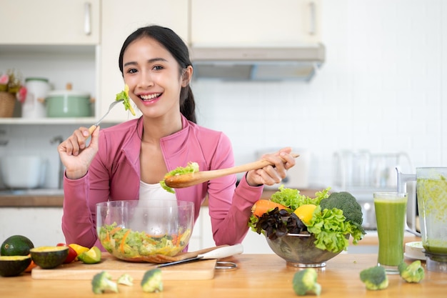 Uma mulher asiática atraente em roupas esportivas desfruta de sua salada saudável em uma mesa de cozinha na cozinha