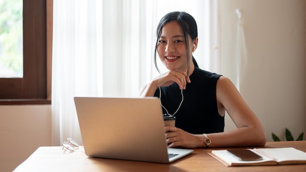 Uma mulher asiática atraente está sorrindo para a câmera enquanto está sentada em um café com seu laptop