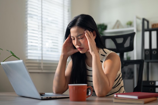 Uma mulher asiática cansada senta-se em frente a um computador lutando contra uma forte dor de cabeça enquanto trabalha