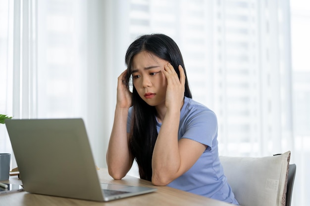 Uma mulher asiática estressada está sentada em uma mesa em seu quarto olhando para a tela do laptop com uma cara de dúvida