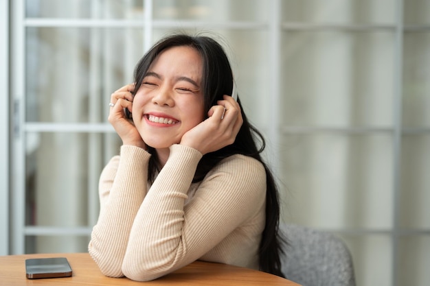 Uma mulher asiática feliz está sentada em uma mesa dentro de casa e desfrutando da música em seus fones de ouvido
