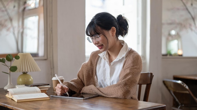 Uma mulher asiática feliz trabalhando remotamente em um café usando seu tablet digital em uma mesa dentro de casa