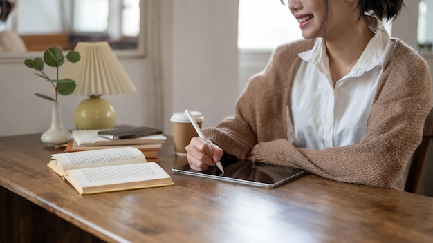 Uma mulher asiática feliz trabalhando remotamente em um café usando seu tablet digital em uma mesa dentro de casa