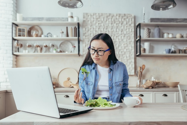 Uma mulher asiática interessada está sentada à mesa da cozinha em casa observa atentamente o online