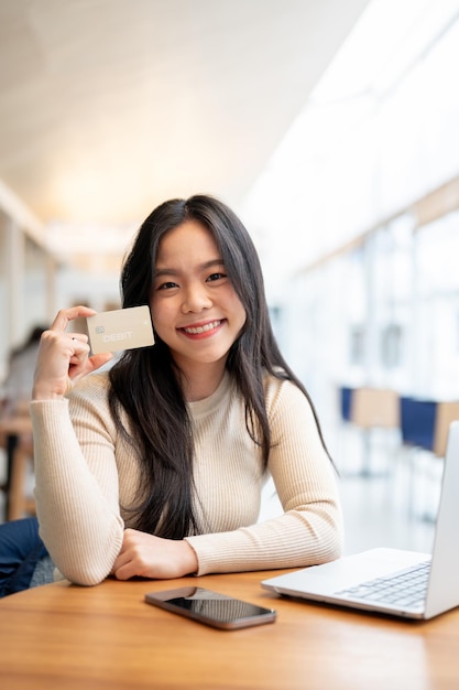 Uma mulher asiática mostrando seu cartão de débito para a câmera enquanto estava sentada em uma mesa em uma cafeteria