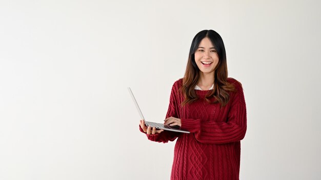 Uma mulher asiática positiva está de pé contra um fundo branco isolado com seu laptop