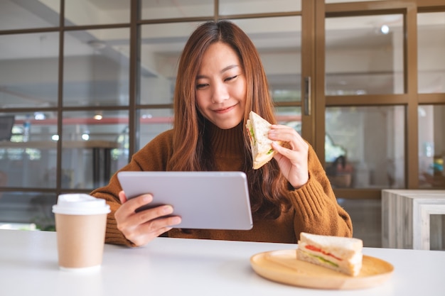 Uma mulher asiática segurando e comendo um sanduíche de trigo integral enquanto usa o tablet pc