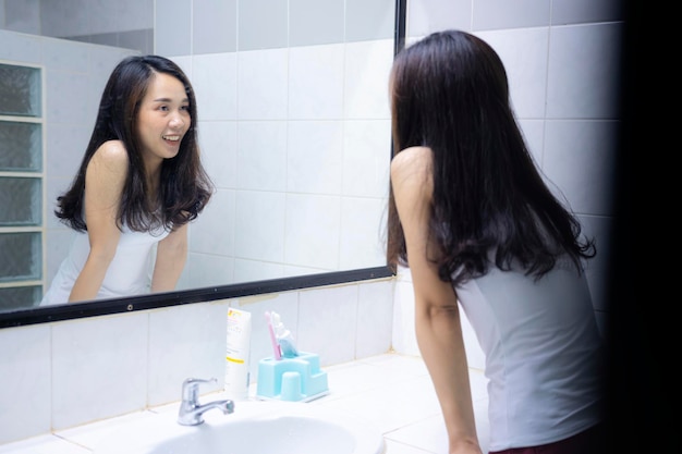 Uma mulher asiática sorrindo para si mesma na frente de um espelho no banheiro em uma nova manhã brilhante