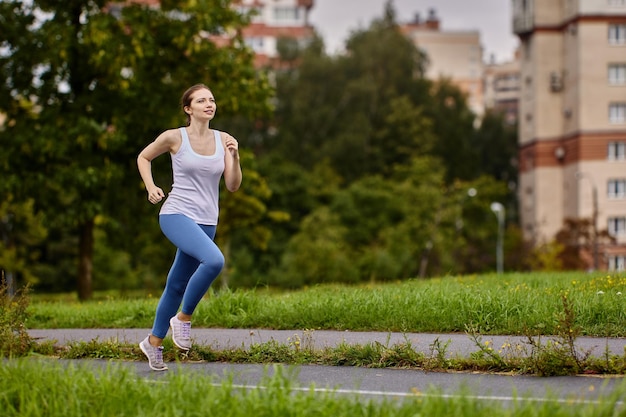 Uma mulher atleta se aquece correndo no parque da cidade de verão perto da área residencial com prédios residenciais de vários apartamentos