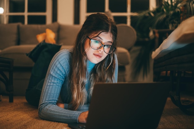 Uma mulher atraente caucasiana e jovem usando óculos usando computador no tapete em casa à noite Linda senhora relaxando navegando na rede interna assistindo vídeos ou trabalhandoxA