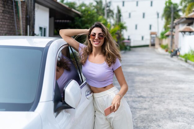 Foto uma mulher atraente e bronzeada sorridente e bem-sucedida em roupas casuais está usando seu telefone inteligente enquanto está em pé