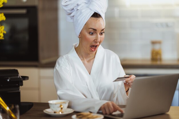 Uma mulher atraente em roupão de banho fazendo compras online em seu laptop enquanto desfruta do café da manhã e relaxa em casa.