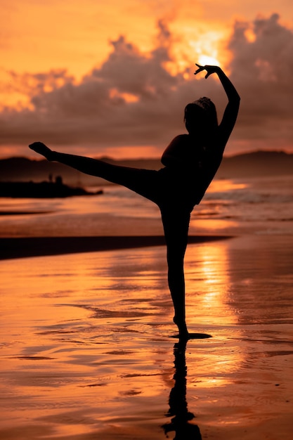 Uma mulher balinesa na forma de uma silhueta executa movimentos de balé com muita habilidade e flexibilidade na praia com as ondas quebrando