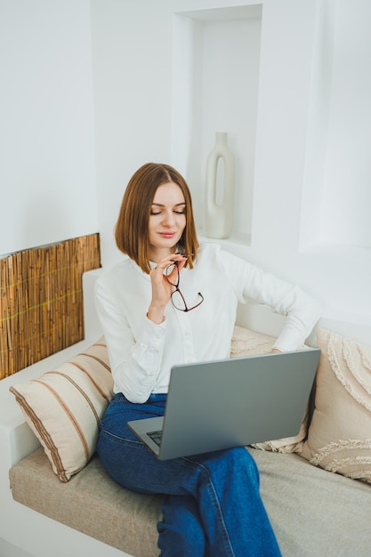 Foto uma mulher bonita de óculos e jeans senta-se em casa e trabalha remotamente em um laptop trabalhe enquanto relaxa em casa