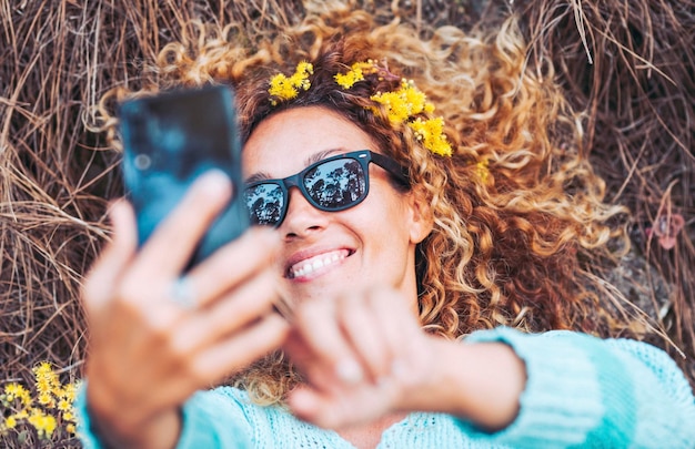 Uma mulher bonita e alegre tirando uma foto de selfie deitada no chão na floresta e usando flores amarelas Pessoas e mídias sociais compartilhando conteúdo Estilo de vida natural e bem-estar