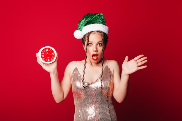 Uma mulher bonita e feliz com um chapéu de Papai Noel está segurando um despertador em um fundo vermelho O conceito da temporada de Natal Natal e ano novo