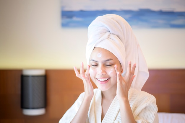 Uma mulher bonita e feliz em um roupão de banho branco, aplicando creme hidratante no rosto no quarto, conceito de cuidados com a pele e tratamento