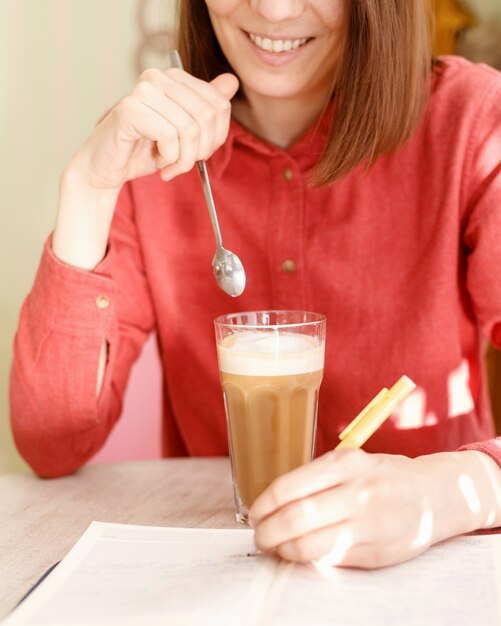 Uma mulher branca com uma camisa rosa sorri e segura uma xícara de café com as mãos em uma cafeteria