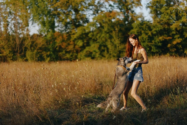 Uma mulher brinca e dança com um cão de raça husky na natureza no outono em um campo de grama e sorri em uma boa noite sob o sol poente