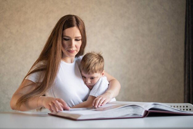 Uma mulher caucasiana de meia-idade abraça um filho pequeno e mostra um álbum de fotos Um menino senta-se no colo de sua mãe e olha para fotografias antigas Lembranças agradáveis Relacionamentos familiares próximos