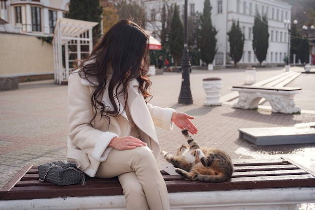 Uma mulher caucasiana despreocupada com roupas bege, apreciando a vista do mar em um dia quente e ventoso. acariciando um gato bonito sentado em um banco nas ruas da cidade.