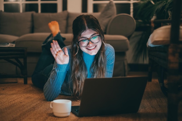 Uma mulher caucasiana e jovem atraente usando óculos usando computador no tapete em casa à noite Linda senhora relaxando navegando na net interna falando em videochamada ou conferênciaxA