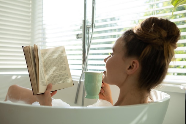 Uma mulher caucasiana passando um tempo em casa, tomando um banho de espuma, lendo um livro, bebendo. Estilo de vida em casa isolando, distanciamento social em quarentena durante a pandemia de coronavírus covid 19.