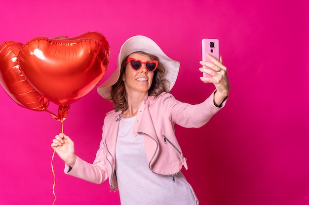 Uma mulher caucasiana se divertindo com um chapéu branco em uma boate com alguns balões de coração tirando uma selfie com o fundo rosa do celular