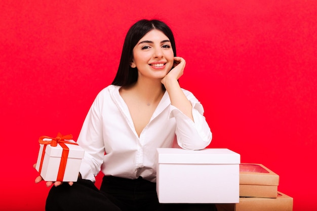 Uma mulher caucasiana sorridente com presentes está sentada em um fundo vermelho no estúdio presentes de Natal em caixas