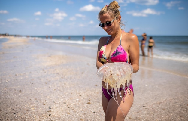 Uma mulher cautelosa e curiosa descontente segura uma água-viva venenosa perigosa morta translúcida em suas mãos e a espalha enquanto está na costa do mar, sob o sol escaldante