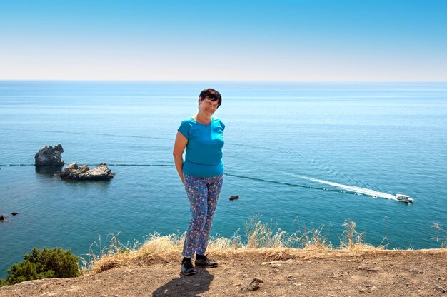 Uma mulher com calças coloridas fica na encosta de uma montanha tendo como pano de fundo o mar