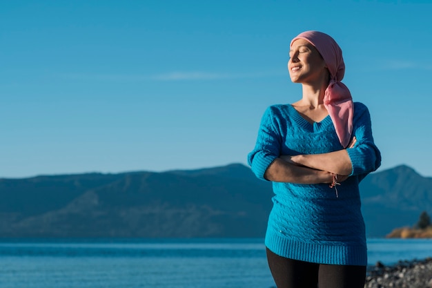 Foto uma mulher com câncer usa um lenço rosa na cabeça e fica com os braços cruzados e sorrindo