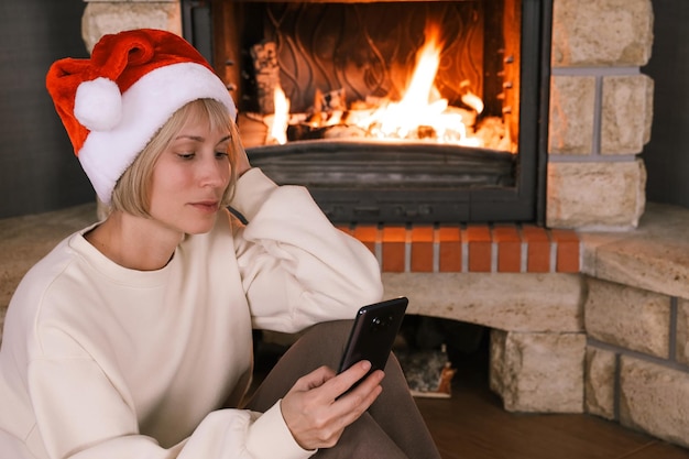 Uma mulher com chapéu de papai noel se comunica ao telefone ao lado da lareira em uma casa aconchegante