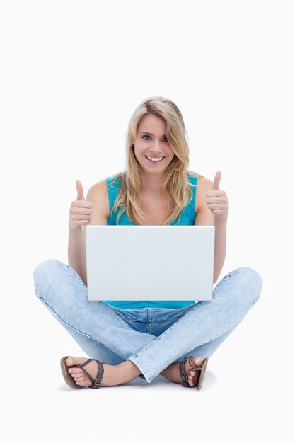 Uma mulher com o polegar está sentada no chão com um laptop