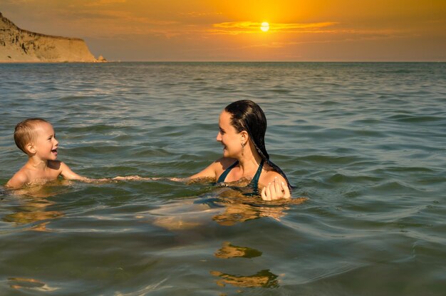 Uma mulher com seu filho no mar contra o fundo do sol.