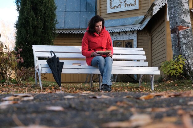 Uma mulher com um casaco vermelho está sentada em um banco branco no parque e lendo um livro de papel.