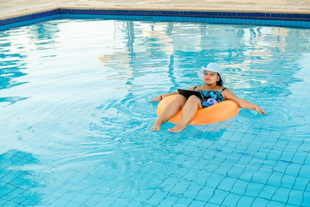 Uma mulher com um chapéu de sol na piscina em um círculo de natação com um laptop se comunica via link de vídeo.
