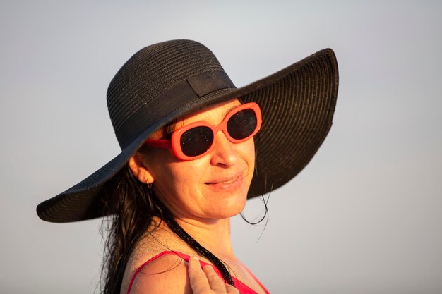 Uma mulher com um chapéu preto de abas largas e óculos de sol está olhando para a câmera e sorrindo