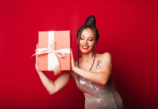 Uma mulher com um chapéu vermelho de Natal com dreadlocks segura um presente em um fundo vermelho Isolar uma mulher em um vestido inteligente ano novo natal