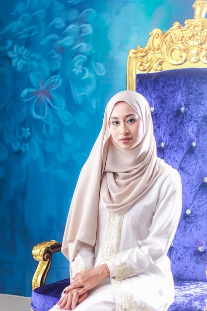 Uma mulher com um hijab branco fica em frente a um trono dourado.