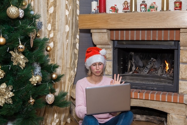 Uma mulher com um laptop com um chapéu de Papai Noel em frente à lareira na sala de estar