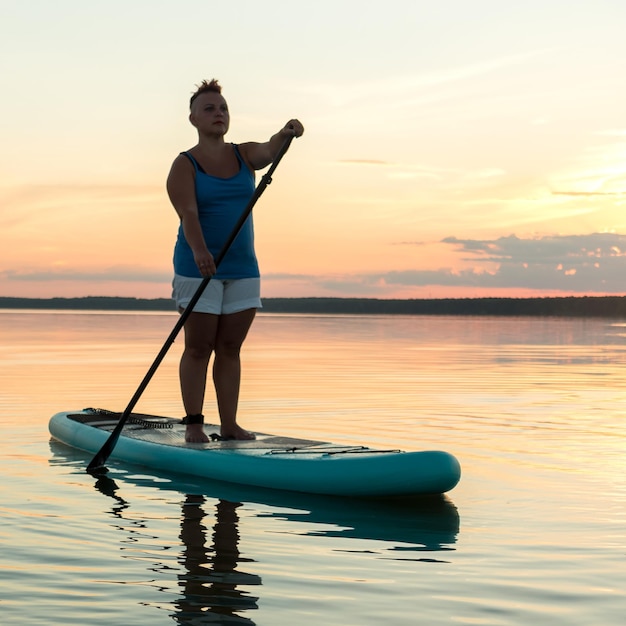 Uma mulher com um moicano em shorts em um sapboard com um remo contra o pano de fundo de um céu brilhante do sol nada no lago à noite