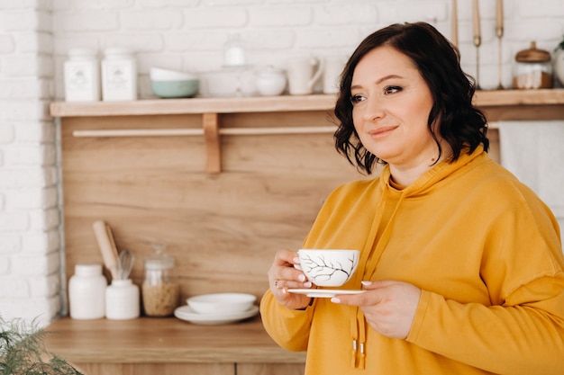 Uma mulher com um moletom laranja bebe café na cozinha de casa.