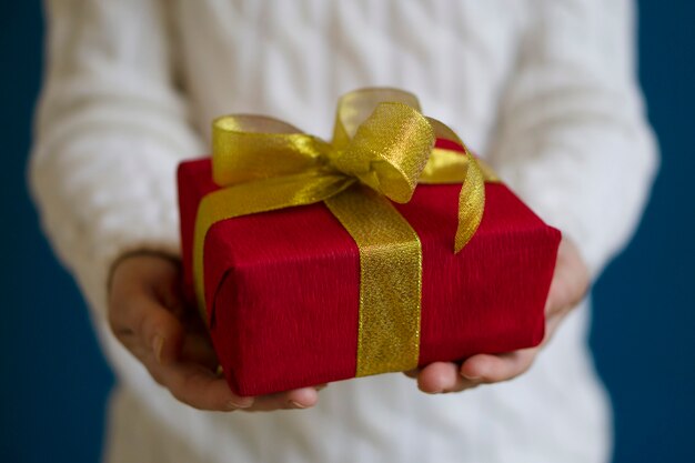 Uma mulher com um suéter branco segura um presente de Natal vermelho sobre um fundo azul. Conceito de presente para o Natal e ano novo.