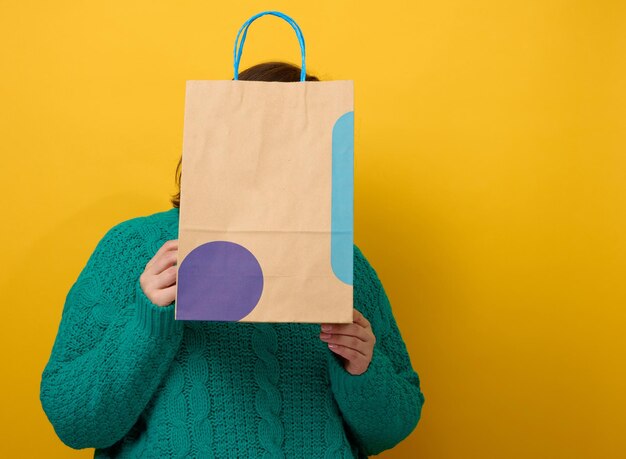 Uma mulher com um suéter segura um saco de papel sobre um fundo amarelo e mostra um gesto semelhante com a mão. compras. venda sazonal