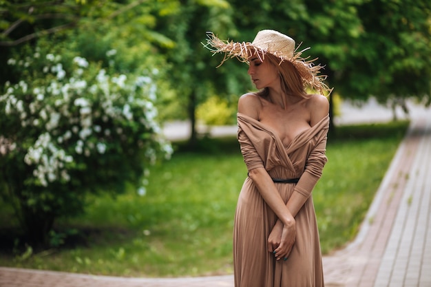 Uma mulher com um vestido longo marrom e um chapéu de palha caminha em um parque de verão