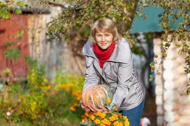 Uma mulher com uma abóbora está no jardim Mulher de meia-idade em jeans e jaqueta no fundo das árvores de outono no quintal