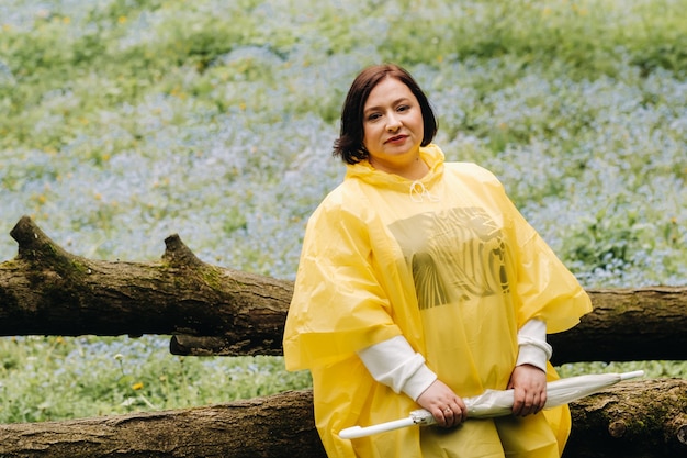 Uma mulher com uma capa de chuva amarela com um guarda-chuva se senta em uma árvore caída na floresta no verão.