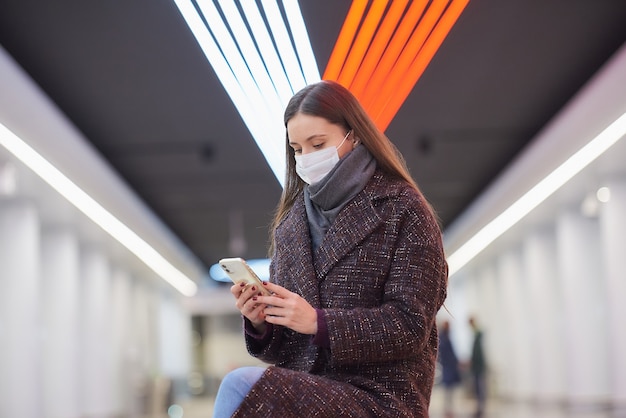 Uma mulher com uma máscara facial de médico está sentada no centro da plataforma do metrô com um smartphone e lendo as notícias